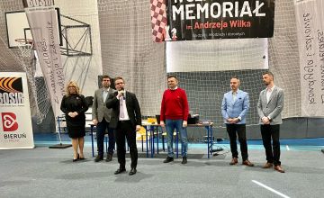 Hala sportowa, w rzędzie stoi 5 osób jest to 4 mężczyzn i 1 kobieta. Kobieta stoi pierwsza z lewej. Krok przed nimi mężczyzna ubrany w garnitur przemawia do mikrofonu. Za wszystkimi znajdują się połączone dwa stoliki szklone, na których znajduje się drukarka oraz laptop. Na ścianie hali zawieszona jest siatka a na niej duży plakat. Z lewej strony plakatu znajduje się grafika z częścią szachownicy oraz pionkami. W jego centralnej części  widnieje napis IV Szachowy Memoriał im. Andrzeja Wilka dla dzieci, młodzieży i dorosłych.